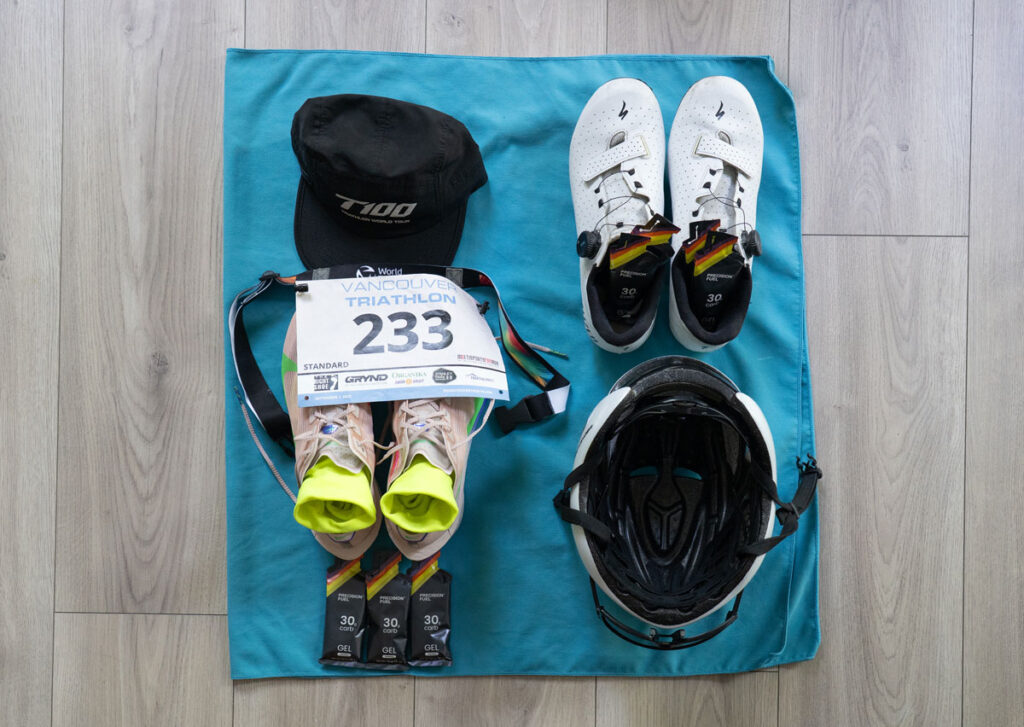 Triathlon transition gear laid out on a teal towel including cycling shoes, helmet, running shoes with socks, race number belt, cap, and energy gels