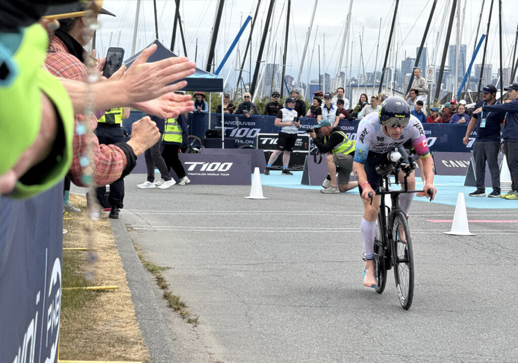 A professional triathlete racing on the bike leg at T100 Vancouver, with spectators cheering along the course